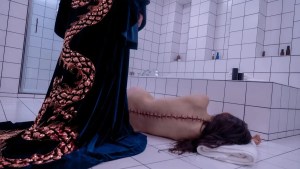 A person in a robe depicting a long snake/dragon-like creature on its back stands over a woman lying on the floor of a bathroom near a shower, her back toward the camera, crude stitches running all the way down her spine.